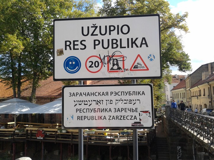 Placa na entrada à República Uzupio com alguns alertas, como a obrigação de sorrir, a velocidade máxima para carros (20km/h) e o do perigo de cair no rio com o carro